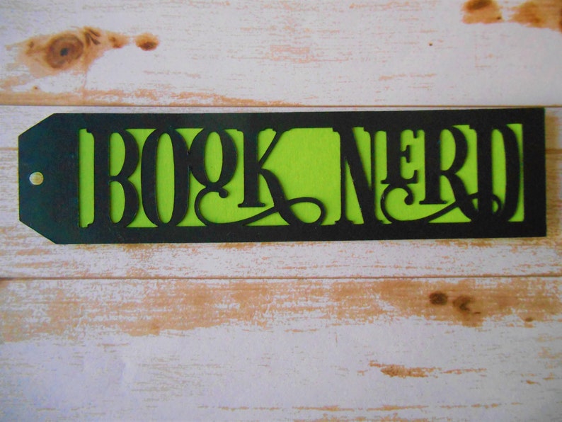 K&ouml;nnte beinhalten: Schwarzer und gr&uuml;ner Lesezeichen mit den ausgeschnittenen Worten "BOOK NERD", die einen gr&uuml;nen Hintergrund freilegen.