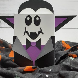 May include: A black and purple papercraft vampire with a white face, big eyes, and fangs. The vampire is wearing a black suit with a purple bow tie. The vampire is sitting on a black and orange fabric with stars.