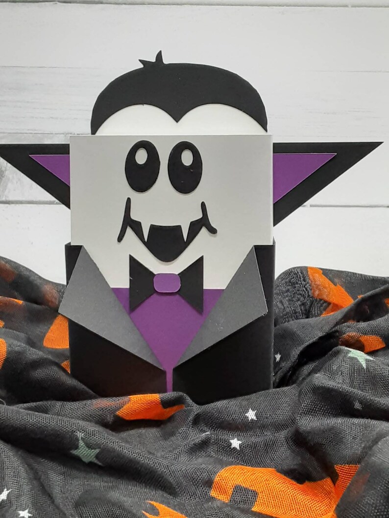 May include: A black and purple papercraft vampire with a white face, big eyes, and fangs. The vampire is wearing a black suit with a purple bow tie. The vampire is sitting on a black and orange fabric with stars.