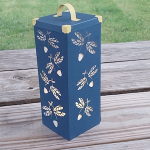 May include: A blue paper lantern with a handle and a cut-out design of acorns and leaves. The lantern has a yellow trim.