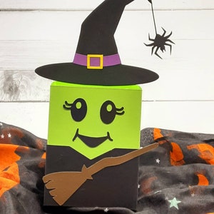 May include: A green witch face Halloween treat box with a black witch hat, a brown broom, and a black spider hanging from the hat.
