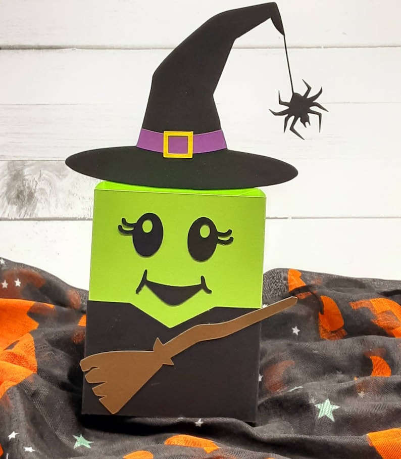 May include: A green witch face Halloween treat box with a black witch hat, a brown broom, and a black spider hanging from the hat.