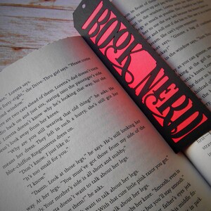 K&ouml;nnte beinhalten: Ein schwarzes und rotes Lesezeichen mit den ausgeschnittenen Worten "Book Nerd". Das Lesezeichen liegt auf einem Buch mit Text &uuml;ber die Beine eines M&auml;dchens.