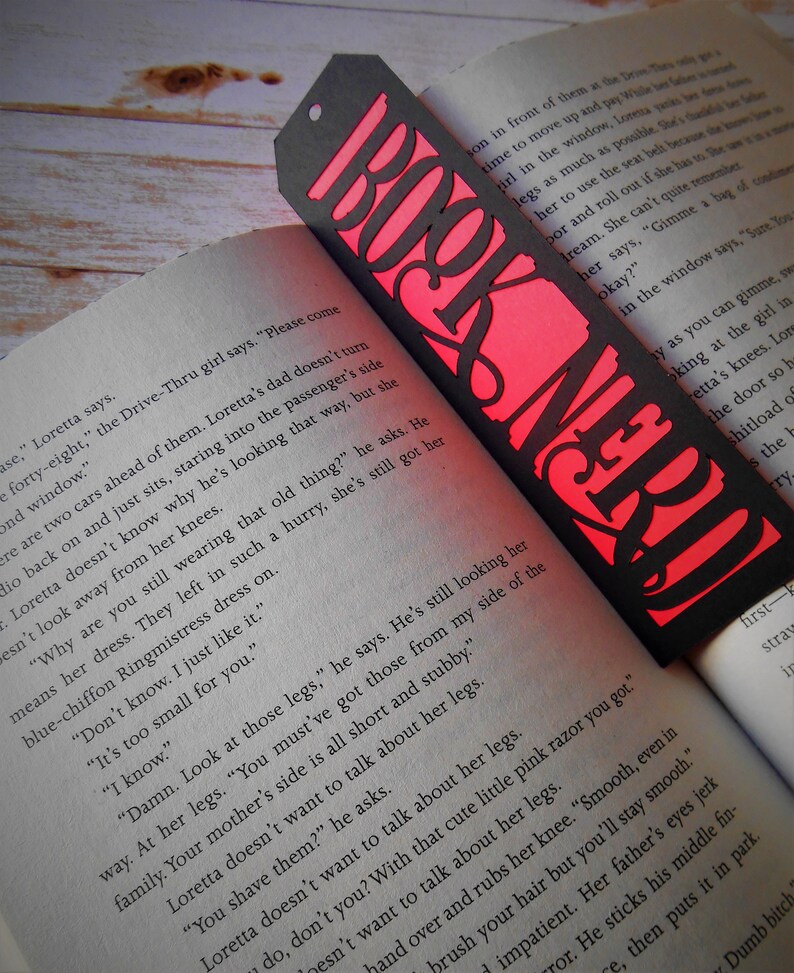 K&ouml;nnte beinhalten: Ein schwarzes und rotes Lesezeichen mit den ausgeschnittenen Worten "Book Nerd". Das Lesezeichen liegt auf einem Buch mit Text &uuml;ber die Beine eines M&auml;dchens.