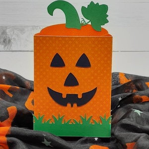 May include: Orange and green paper pumpkin treat box with a jack-o'-lantern face. The box is sitting on a black and orange fabric with stars.