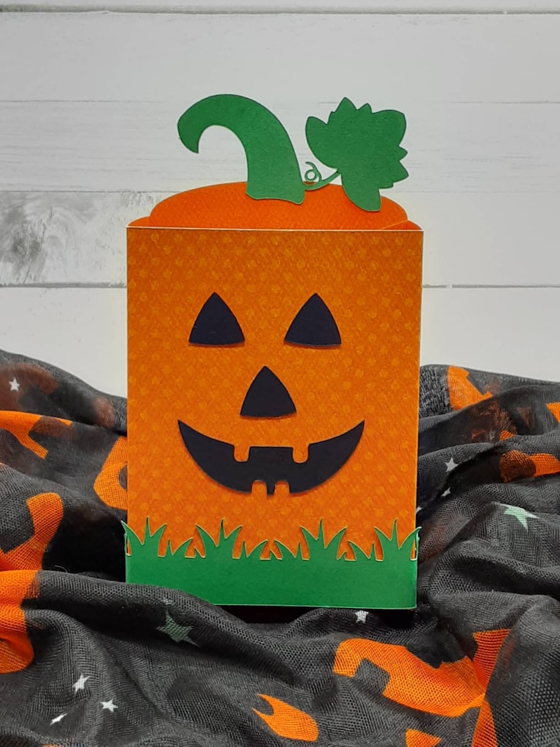 May include: Orange and green paper pumpkin treat box with a jack-o'-lantern face. The box is sitting on a black and orange fabric with stars.