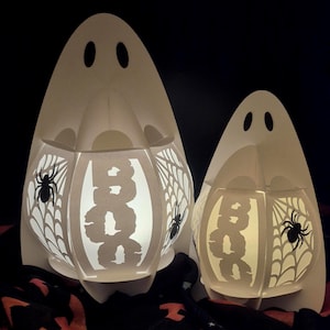 Könnte beinhalten: Zwei weiße Geisterlaternen aus Papier mit ausgeschnittenen Designs. Die Laternen haben das Wort "BOO" auf der Vorderseite ausgeschnitten und Spinnweben und Spinnen an den Seiten. Die Laternen sind von innen beleuchtet und schaffen eine gruselige Halloween-Dekoration.
