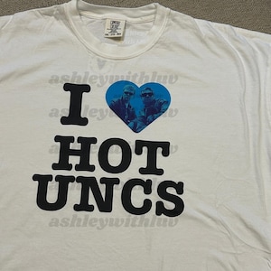 Puede incluir: Camiseta blanca con el texto "I HOT UNCS" en letras negras. Un corazón azul contiene una foto de dos hombres con gafas de sol. La marca "Comfort Colors" es visible en la etiqueta.