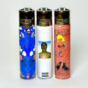 May include: Three cylindrical Clipper lighters with metallic tops. The left lighter is blue with a repeating cartoon mouse pattern. The center lighter is white with a portrait of a person and the text Parental Advisory. The right lighter is red with a graphic of a person and a paisley pattern.