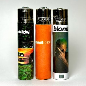 May include: Three cylindrical Clipper lighters with metallic tops. The left lighter features a car and the text 'nostalgia, ULTRA'. The center lighter is solid orange with 'channel ORANGE' text. The right lighter shows a person and 'blond' text.