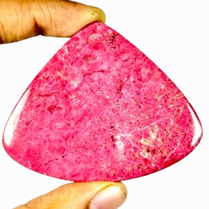 May include: A triangular, polished gemstone in a vibrant pink hue. The stone has a textured surface with subtle variations in colour and small, light-coloured veins. The gemstone is held between two fingers against a white background.