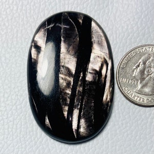 May include: A black and silver oval cabochon gemstone with a striated pattern. The gemstone is approximately the size of a US quarter.