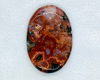 May include: An oval-shaped cabochon gemstone featuring a complex pattern of swirling colors including deep rust red, burnt orange, dark brown, and patches of muted lavender and black, with a smooth, polished, and reflective surface finish.