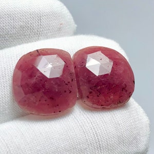 May include: Two faceted, oval-shaped gemstones with a deep pink hue. The gems have a polished, reflective surface with multiple facets, and are set against a white background. The gems are likely rubies or similar precious stones.