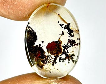 Könnte beinhalten: Ovaler, durchscheinender, weißer Achat-Cabochon mit schwarzen und roten dendritischen Einschlüssen. Der Stein hat ein natürliches, organisches Muster.