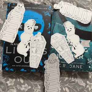 May include: A collection of five black and white die-cut stickers featuring illustrations of ghosts, a coffin-shaped bookshelf, and creatures perched atop stacks of books, arranged on top of two dark-coloured books with visible text and floral patterns.