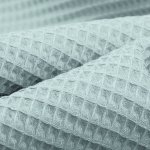 May include: Close-up of a light blue waffle-weave fabric. The fabric is folded, showing the textured pattern of small, evenly spaced squares. The material appears soft and pliable, with subtle variations in tone.