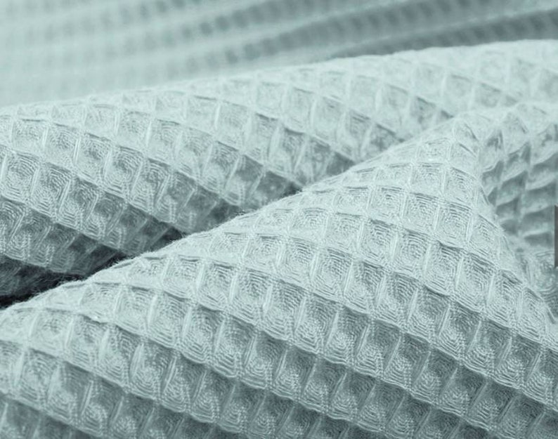 May include: Close-up of a light blue waffle-weave fabric. The fabric is folded, showing the textured pattern of small, evenly spaced squares. The material appears soft and pliable, with subtle variations in tone.