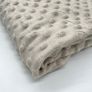 Könnte beinhalten: Eine gefaltete, beige Decke mit einem strukturierten, gepunkteten Muster. Der Plüschstoff wirkt weich und einladend, mit einem dezenten Glanz. Die Decke ist gefaltet und zeigt die Textur und Farbe.