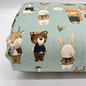 May include: A blue fabric with a repeating pattern of various animals wearing clothing. The animals include a deer, a bear, a rabbit, a fox, and a goose. The animals are all wearing different outfits, including vests, scarves, and hats.
