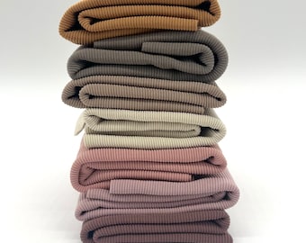 May include: A pile of folded ribbed fabric in a range of neutral tones, such as tan, grey, beige, and pink and brown hues. The fabric has a textured, corduroy-like look, and is neatly stacked against a white backdrop.