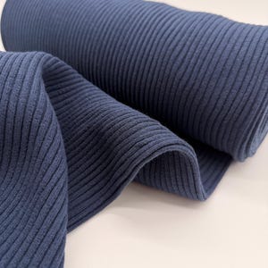 Könnte beinhalten: Eine Rolle marineblauen Rippstrickstoffs. Der Stoff ist eng gerollt, mit einem Teil ausgerollt und drapiert, wodurch die vertikale Rippstruktur zur Geltung kommt. Der Stoff hat ein weiches, mattes Aussehen, geeignet für Bekleidung oder Heimdekorationsprojekte.