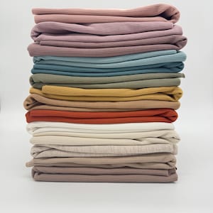Könnte beinhalten: Ein Stapel von 12 verschiedenen, gerippten Stoffproben in verschiedenen Farben. Die Farben sind staubiges Rosa, hellblau, olivgrün, senfgelb, gebranntes Orange, weiß, beige, hellbraun und dunkelbraun.