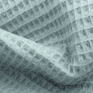 May include: Close-up of a light blue waffle-weave fabric. The fabric has a textured, grid-like pattern, with a soft, slightly crumpled appearance. The weave creates a subtle interplay of light and shadow, highlighting the texture. The fabric appears to be a towel or blanket.