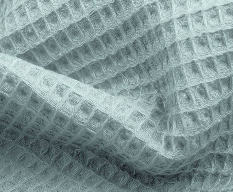 May include: Close-up of a light blue waffle-weave fabric. The fabric has a textured, grid-like pattern, with a soft, slightly crumpled appearance. The weave creates a subtle interplay of light and shadow, highlighting the texture. The fabric appears to be a towel or blanket.