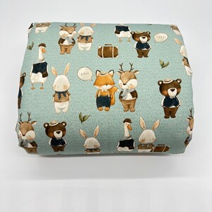 May include: A light blue fabric with a repeating pattern of various animals wearing hats and scarves. The animals include a fox, deer, rabbit, goose, and bear. The fabric has a subtle textured pattern.