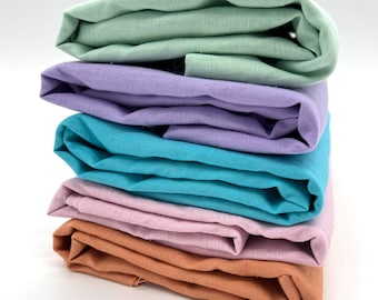 Könnte beinhalten: Ein Stapel gefalteter Stoffe in verschiedenen Pastellfarben. Die Farben umfassen Hellgrün, Lavendel, Türkis, Rosa und ein gedämpftes Orangebraun. Der Stoff scheint Leinen oder ein ähnliches strukturiertes Material zu sein.