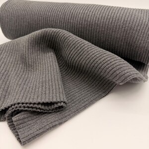 Könnte beinhalten: Eine Rolle grauer Rippstrickstoff, mit einem gefalteten Abschnitt im Vordergrund. Der Stoff hat vertikale Rippen und eine weiche Textur. Die Farbe ist ein mittleres Grau, geeignet für verschiedene Nähprojekte.