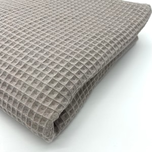 May include: A close-up of a folded waffle weave fabric in a light brown color. The fabric has a textured, waffle-like pattern.