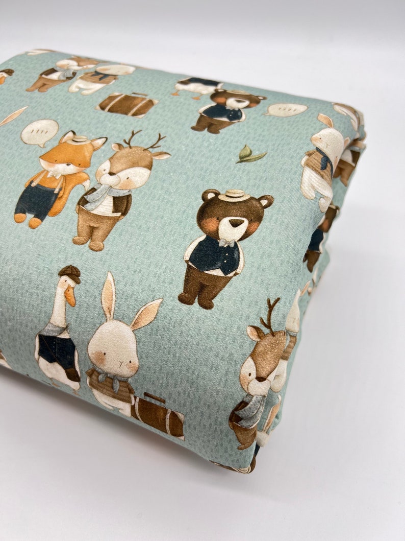May include: A light blue fabric with a repeating pattern of various animals wearing hats and carrying suitcases. The animals include a fox, a deer, a bear, a rabbit, and a goose. The fabric has a subtle texture.
