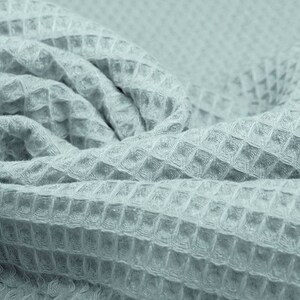 May include: Close-up of a light blue waffle-weave fabric. The fabric is folded and draped, showcasing its textured surface. The weave pattern creates a grid-like appearance, adding visual interest. The fabric appears soft and pliable, suitable for various textile projects.