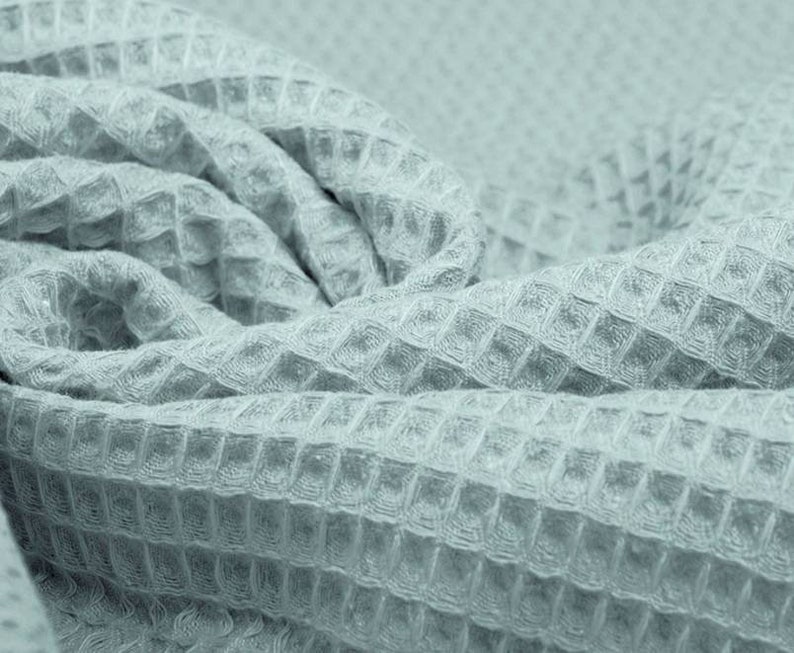 May include: Close-up of a light blue waffle-weave fabric. The fabric is folded and draped, showcasing its textured surface. The weave pattern creates a grid-like appearance, adding visual interest. The fabric appears soft and pliable, suitable for various textile projects.