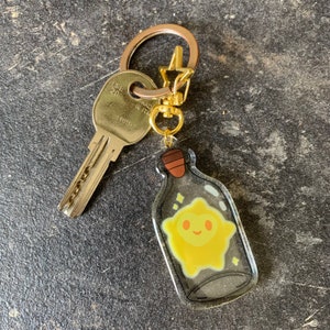 May include: A gold key chain with a clear bottle charm. The bottle contains a yellow star with a smiling face. The star is surrounded by glitter.