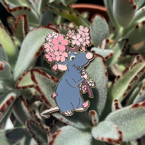 Op de afbeelding: Een blauwe rattenemaille pin met roze oren en voeten, die een tak met roze kersenbloesems vasthoudt. De pin heeft een gouden omtrek en staat tegen een achtergrond van groene succulentenbladeren.