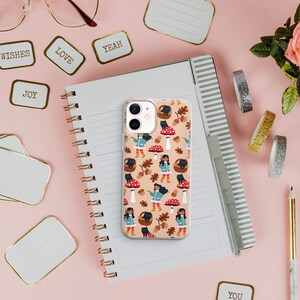 May include: A white iPhone case featuring a colourful pattern of mushrooms, leaves, and a girl wearing a blue jacket and red boots. The case is on a notebook with a spiral binding.