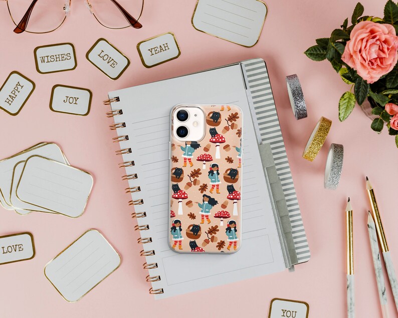 May include: A white iPhone case featuring a colourful pattern of mushrooms, leaves, and a girl wearing a blue jacket and red boots. The case is on a notebook with a spiral binding.