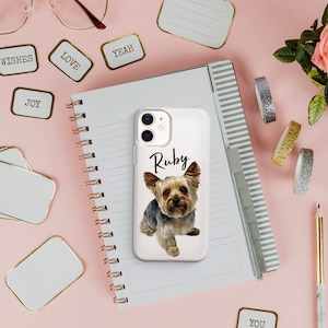 Pu&ograve; includere: Custodia trasparente per telefono con un cane Yorkshire Terrier e il nome "Ruby". La custodia &egrave; su una superficie rosa con un taccuino, penne ed etichette decorative. Sono visibili anche una rosa e nastro glitterato.