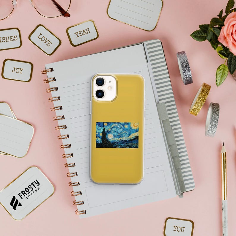 May include: A mustard yellow smartphone case featuring a rectangular print of Vincent van Gogh's The Starry Night painting. The case has a dual-camera cutout at the top left and is positioned on a spiral-bound notebook with lined pages.