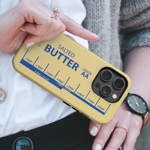 May include: A yellow phone case with a butter-themed design. The case features the words "SALTED BUTTER" in blue, along with measurement markings in tablespoons and cups. The case has a camera cutout and is held by a person.
