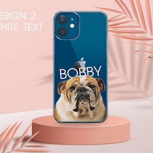 Pu&ograve; includere: Custodia trasparente per telefono con un ritratto di bulldog e il nome "BOBBY" in bianco. La custodia &egrave; su un piedistallo rosa con sfondo rosa chiaro. Il testo "DESIGN 2 WHITE TEXT" &egrave; nell'angolo in alto a sinistra.