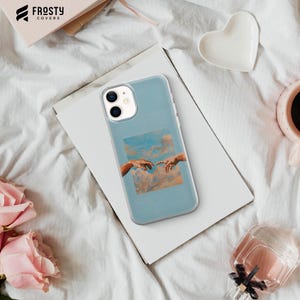 May include: A light blue smartphone case featuring a central square graphic of the Creation of Adam painting, showing two hands reaching toward each other against a backdrop of soft, orange-toned clouds and a light blue sky.