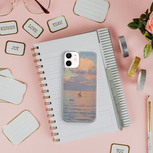 May include: A clear phone case with a sunset and sailboat image on a pink background with a notebook, pencils, and decorative tape.