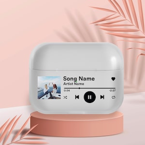 May include: White Airpods case with a custom design featuring a photo of two people on a boat, the text "Song Name", "Artist Name", and a music player control bar with play, pause, and skip buttons.