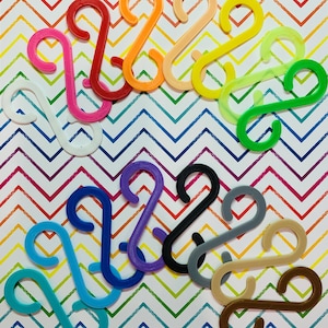 May include: A collection of colorful S-shaped hooks in various shades, including red, orange, yellow, green, blue, purple, black, gray, beige, and brown. The hooks are arranged on a vibrant, multicolored chevron patterned background.