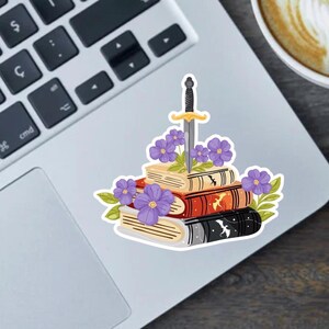 Pode incluir: Um adesivo com uma pilha de livros com uma espada atravessando-os, adornada com flores roxas e folhas verdes. Os livros são em várias cores, incluindo bege, vermelho e preto. O adesivo está em um laptop.