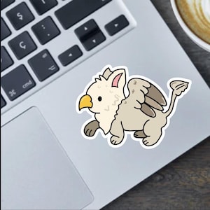 May include: A sticker featuring a cartoon griffin with a cream-colored body, tan wings, and a yellow beak. The griffin is depicted in a walking pose, with a black outline. The sticker is placed on a laptop.